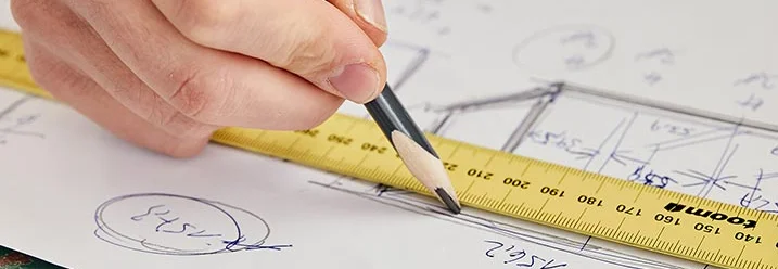 Eine Hand zeichnet mit einem Bleistift auf einem Blatt Papier mit Bauplänen. Ein Zollstock dient als Lineal.